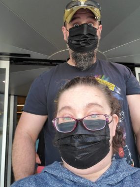 An adult man and woman wearing black disposable face masks exit a medical building. The woman is wearing a blue hoodie and glasses and has her hair tied back. She appears to be seated in a wheelchair, and her husband is standing behind her. He's wearing a green baseball cap, sunglasses, and a blue T-shirt and has a beard sticking out beneath his face mask.