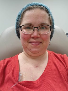 The photo shows a middle-aged Caucasian female with glasses, wearing a red shirt and teal cap, with a medical IV tube poking out of the neck of her shirt. She is receiving IVIG therapy.
