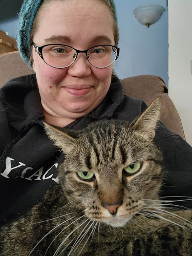 An adult white female with glasses and a beanie hat sits for a closeup selfie while an adult tabby cat sits on her lap. The cat is looking toward the camera and down, with a grumpy-looking expression that makes tabby cats so adorable. 