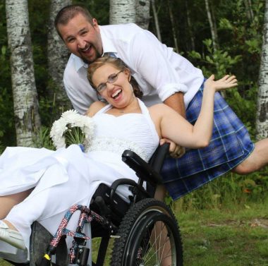 This photo shows a smiling woman with short, blondish-brown hair and glasses in a white wedding dress; she sits in a wheelchair and holds white flowers in one hand while the other hand is raised. The chair's front tires are raised off the ground by a smiling man behind her, who has close-cropped, dark brown hair and a goatee. He wears a white shirt and a blue checked kilt with thin white lines. They are outdoors, and a stand of trees is behind them. 