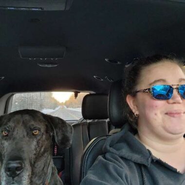 An adult female with sunglasses, sits in a van next to her dog, a black Great Dane. It is a frontal view, from the perspective of the windshield looking back, and past the woman and the dog, a long road stretches off into the horizon, where the sun is either rising or setting. 