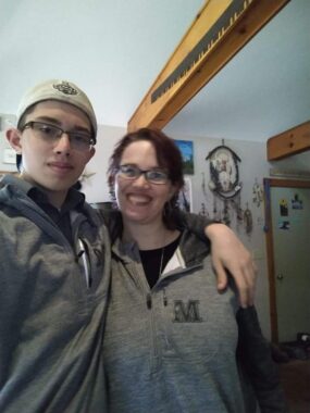 A mom and her son stand next to each other, with the son's arm around her shoulder, and smile for a photo. They're both wearing gray hoodies and glasses.