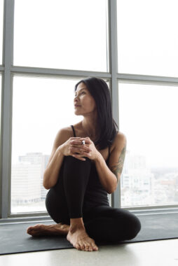 A woman sits on the floor in front of a large picture window in a large city. She is barefoot and wearing yoga clothes. She looks to her right, has her right knee raised, and her hands are clasped on her knee. 