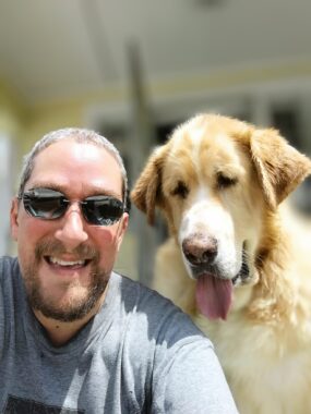 A man with a goatee and sunglasses smiles for a photo with his dog Carmel.