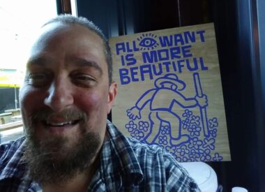 A man wearing a blue flannel shirt smiles in front of a wooden sign that reads "All I want is more beautiful" in blue, with an image of a man hiking through flowers.