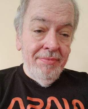 A close-up photo shows a man from the chest up. He has white hair, a mustache, and a short beard, and is wearing a black T-shirt with an orange logo.