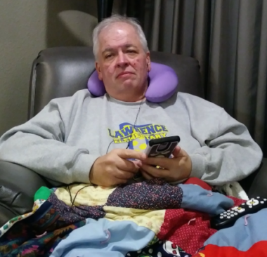 A man wearing a gray sweater sits in what appears to be an infusion chair. He has a purple neck pillow behind his head, a multicolored quilt on his lap, and he's holding his phone in front of him.