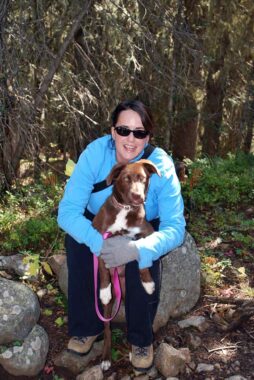 A woman sits on a rock while hugging her dog to her chest. She's wearing a long-sleeve blue top, sunglasses, and hiking boots and pants. The dog is brown and white and looking at the camera. They're on a hiking trail near Nederland, Colorado.