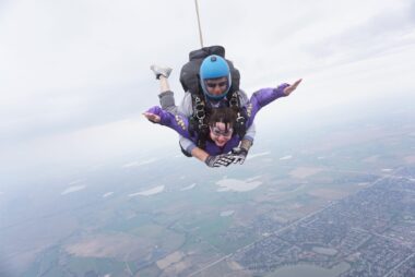 A photo taken midair of a woman skydiving while strapped to an instructor. She's holding her arms out to the sides and is wearing a purple shirt and goggles. They are falling above Longmont, Colorado.