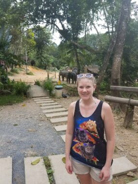 A young woman in a tank top and shorts stands on a path on a horse farm in the tropical woods. 