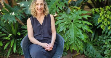 A woman with blond curly hair is smiling and seated on a blue chair outside in front of some lush greenery.