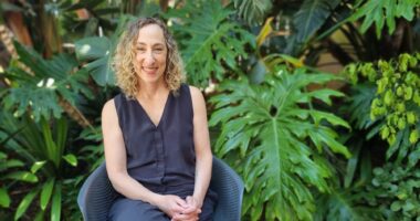 A woman with blond curly hair is smiling and seated on a blue chair outside in front of some lush greenery.