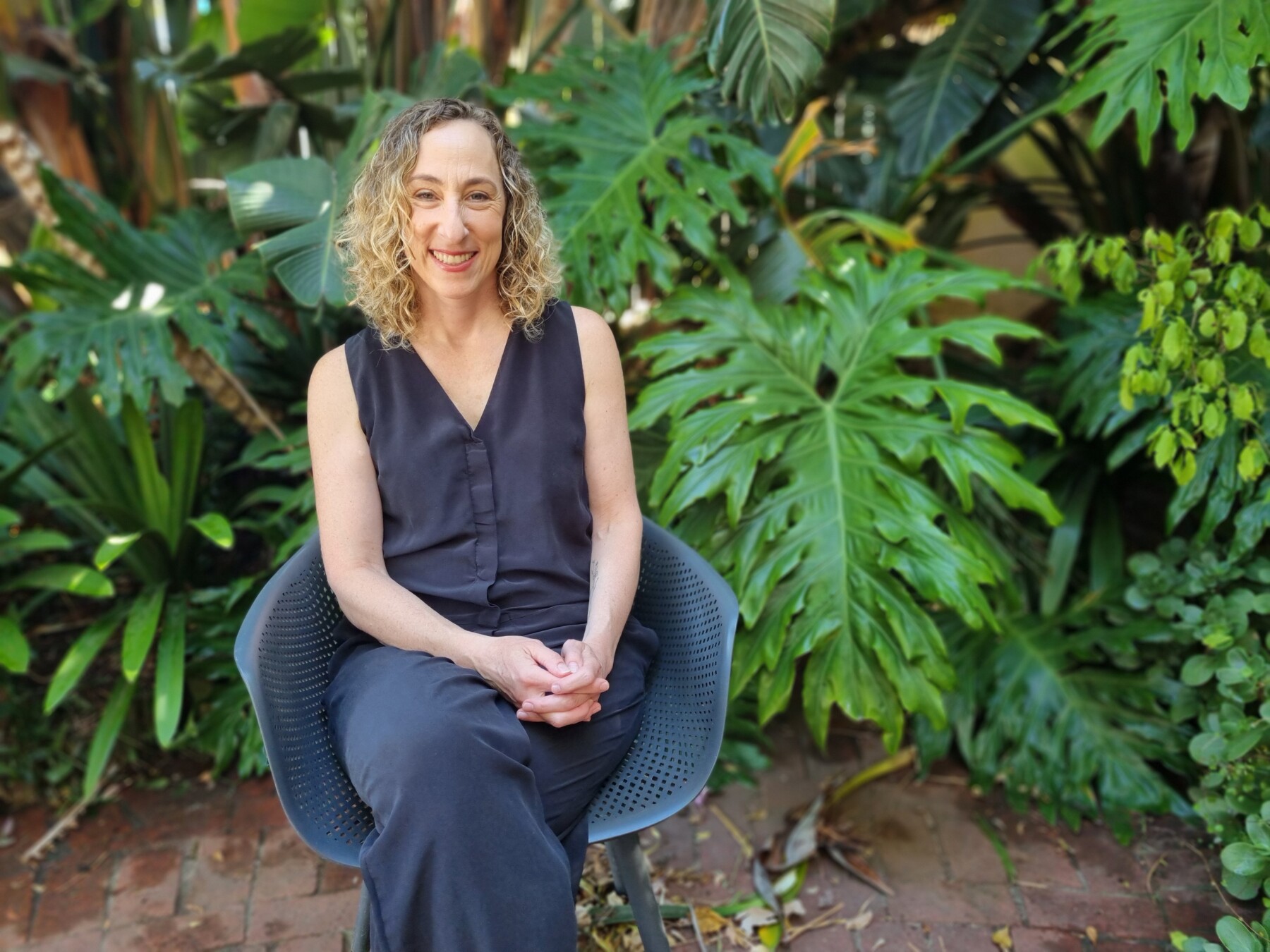 A woman with blond curly hair is smiling and seated on a blue chair outside in front of some lush greenery.