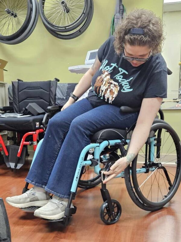 A woman with frizzy brown hair sits in a light teal wheelchair, examining the functions by the front wheel. She wears a black T-shirt with a central design in pale green and tan, blue jeans, and white and gray shoes.