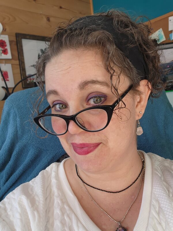 A close-up of a woman looking at the camera on her computer. She looks over a pair of glasses resting on the ridge of her nose and has her head tilted to the right. 