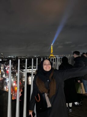 Una joven con un abrigo negro abre los brazos y sonríe en lo alto del Arco de Triunfo de París. Como es de noche, puedes ver las luces de la ciudad y la Torre Eiffel iluminada detrás de ella.