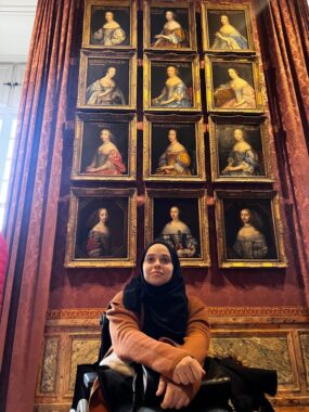 Una mujer sentada en una silla de ruedas frente a un retrato en el Palacio de Versalles. Todas las pinturas representan mujeres, muchas de las cuales parecen pertenecer a la realeza.