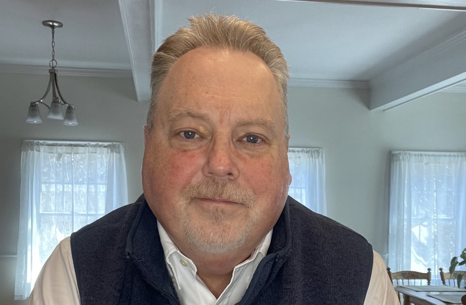 A face shot of a man sitting in his living room, taken from the camera on a computer.