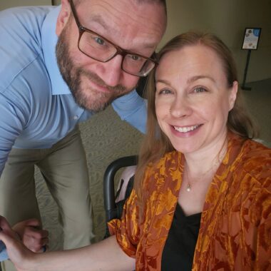 A woman smiles for a photo while seated in her power wheelchair. Her husband, who's standing next to her, leans down so his face is in the frame.