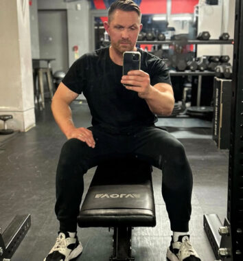 A man in all black gym clothes sits on a weight bench and takes a selfie in a mirror at the gym.