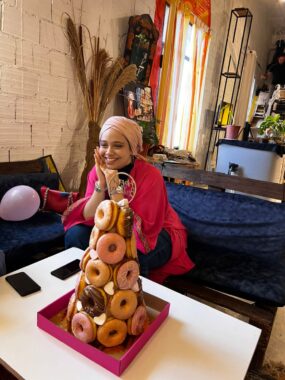 A young woman dressed in a hot pink shawl and a light pink hijab smiles with her hands pressed against her right cheek. She's sitting in front of a low table with a donut tower, arranged in the style of a croquembouche.