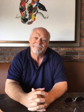 An older man in a navy blue shirt smiles softly at the camera with his hands clasped in front of him. He looks like he might be sitting at a booth in a restaurant.