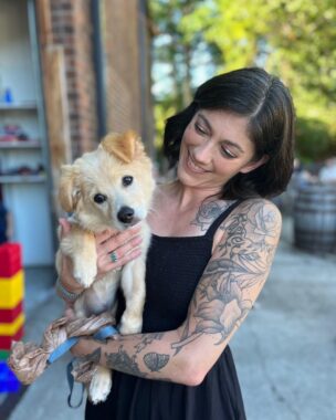 A woman stands outside next to a brick building holding up her dog. She has shoulder-length brown hair and tattoos on her left arm. The dog is looking at the camera, and the woman is smiling down at her dog.