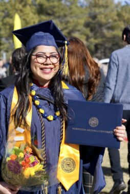 A young woman wearing graduation attire smiles for a photo. She has on a blue cap and gown and a gold stole, and is holding up her diploma in one hand and a bouquet of flowers in the other. She's outside, with tall trees behind her, and it looks to be a sunny day.