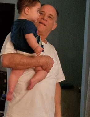 An older man smiles while holding up his young grandson with one arm.