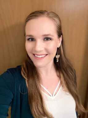 A headshot depicts a woman with long hair smiling at the camera. She's wearing a white blouse under a teal cardigan with long gold earrings and standing against a wooden door.