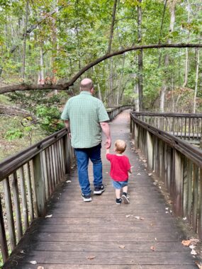 A photo taken from behind shows a balding man with a green shirt and blue jeans walking hand-in-hand with a small boy in a red T-shirt and shorts. They're walking on a wooden nature pathway through the woods.