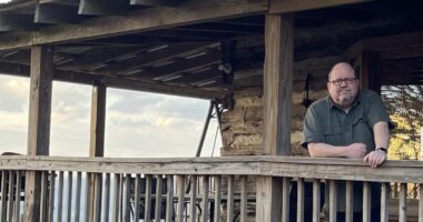 A man with glasses stands on a large, wooden porch or deck, indicating he's in some type of natural setting, like a state park. He's leaning on a wooden rail with his right arm and looking directly into the camera.