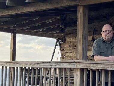 A man with glasses stands on a large, wooden porch or deck, indicating he's in some type of natural setting, like a state park. He's leaning on a wooden rail with his right arm and looking directly into the camera.