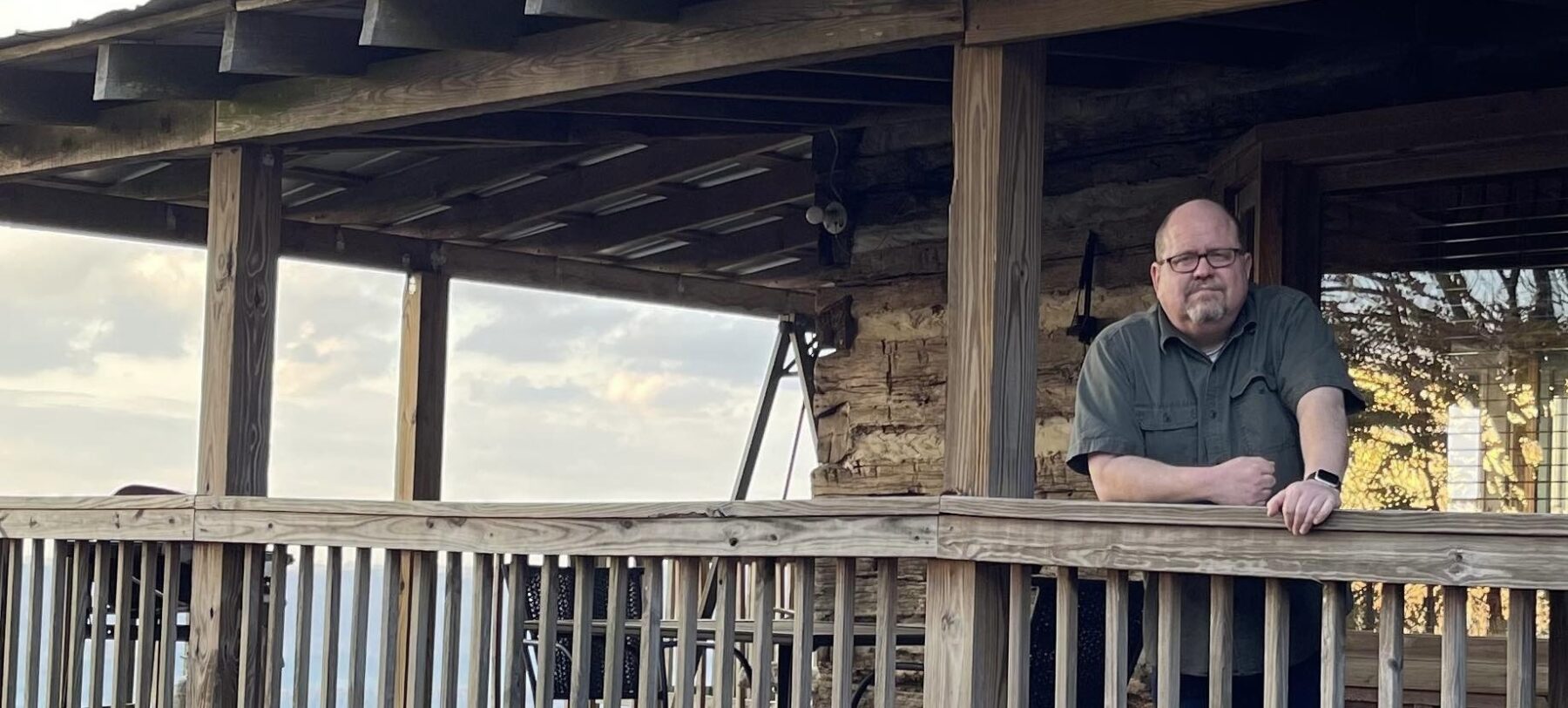 A man with glasses stands on a large, wooden porch or deck, indicating he's in some type of natural setting, like a state park. He's leaning on a wooden rail with his right arm and looking directly into the camera.
