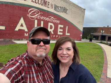 A man and woman pose for the camera in front of a large building with a mural painted on the side that says, "Welcome to historic downtown Laurel, Mississippi."