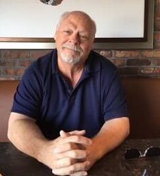 A nearly balding man with a slight smile, a blue polo shirt, and a gray goatee and mustache sits at a table of what appears to be a booth, with a brick wall and the bottom part of largely white painting behind him. His hands are folded in front of him on the table, with a pair of sunglasses nearby.