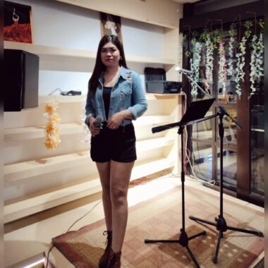 A young woman stands on a small performance stage next to a music stand and a microphone stand. She holds a microphone in her right hand. 
