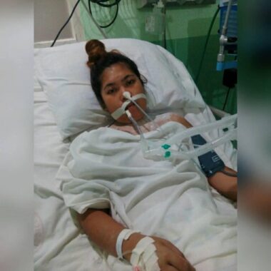 A woman lies in a hospital bed and stares forlornly into the camera. She has a multitude of medical devices hooked up to her, including tubes in her nose and mouth, and various lines to her hand and arm.