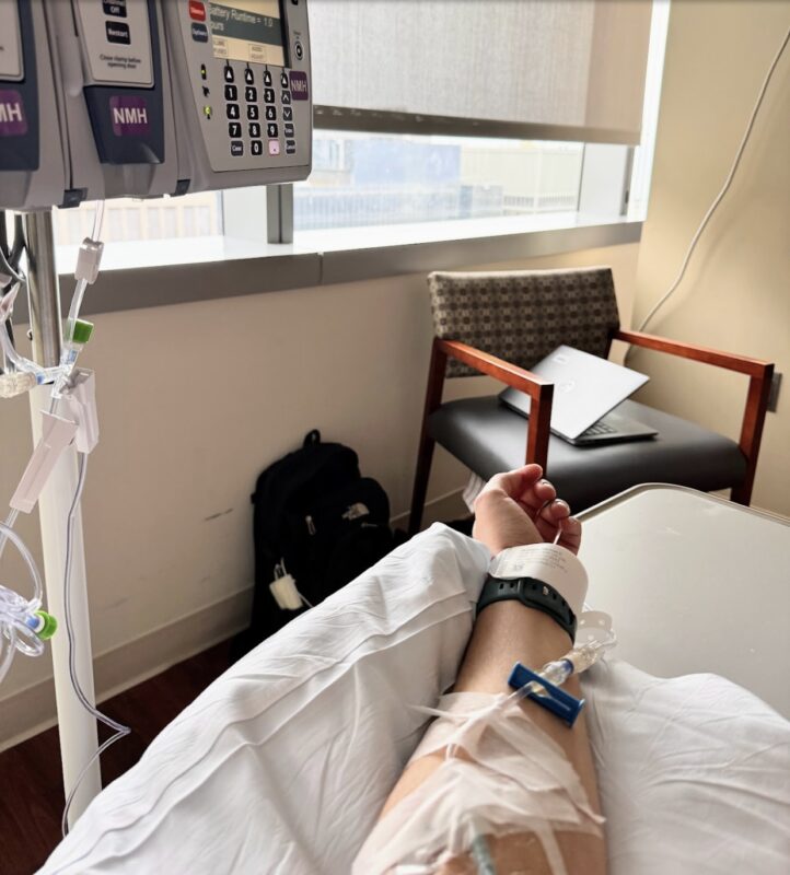 A hospital room setting showing someone's left arm extended out to the edge of a hospital bed and connected to an IV drip. The IV machine is visible to the upper left, and a hospital window is straight ahead, with the shade drawn mostly closed. The arm, bed, and a chair are at weird angles, creating visual discomfort.