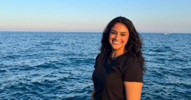 A woman stands slightly angled to the camera, in front of a large, blue body of water (likely Lake Michigan in Chicago_).