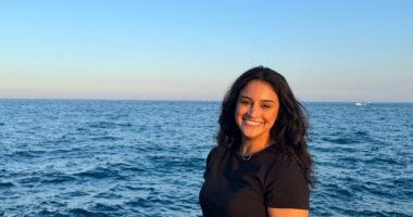 A woman stands slightly angled to the camera, in front of a large, blue body of water (likely Lake Michigan in Chicago_).