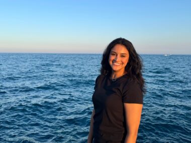 A woman stands slightly angled to the camera, in front of a large, blue body of water (likely Lake Michigan in Chicago_).
