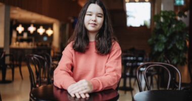 A young woman sits at a small round table with her hands folded in front of her.
