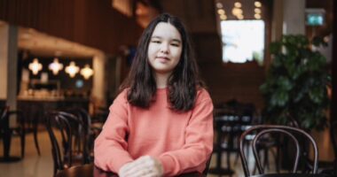 A young woman sits at a small round table with her hands folded in front of her.