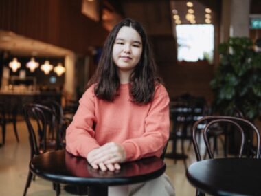 A young woman sits at a small round table with her hands folded in front of her.