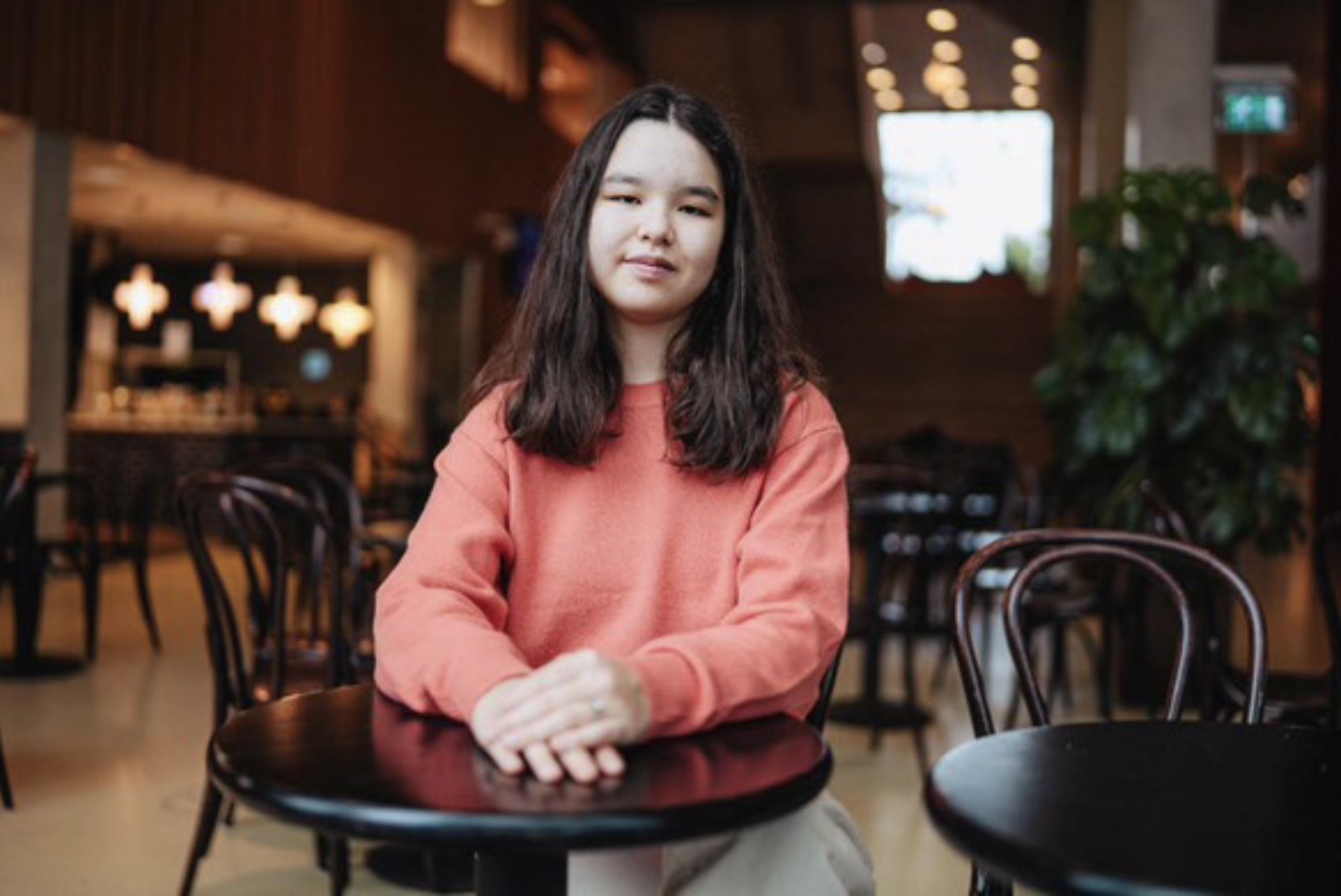 A young woman sits at a small round table with her hands folded in front of her.