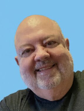 A close-up photo shows a man smiling against a blue background. It kinda looks like a driver's license photo. 