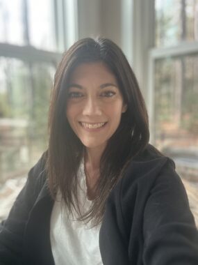 A woman with dark hair is pictured smiling in front of large windows.