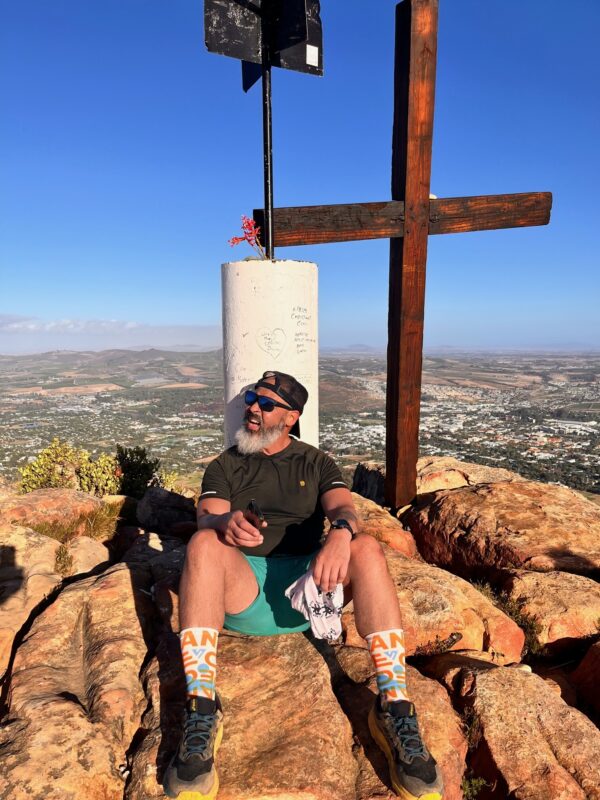 A man with a black backward baseball cap, black sunglasses, and a black T-shirt and green shorts looks off to the right as he sits in the sun atop rocks. Behind him is a large wooden cross. In the background, in a valley below, is a large, flat, urban plain. 
