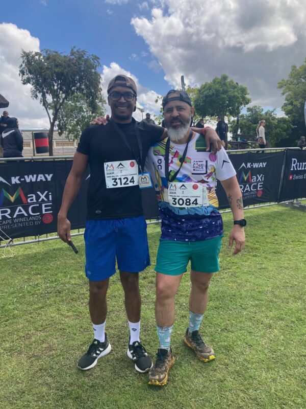 Two men, both wearing backward baseball caps and running clothes, smile and embrace in a side hug at the finish line of a race. 